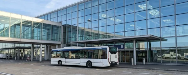Une navette blanche stationnée devant l'architecture vitrée moderne du terminal de l'aéroport Charles de Gaulle, sans passagers visibles, sous un éclairage naturel de matinée