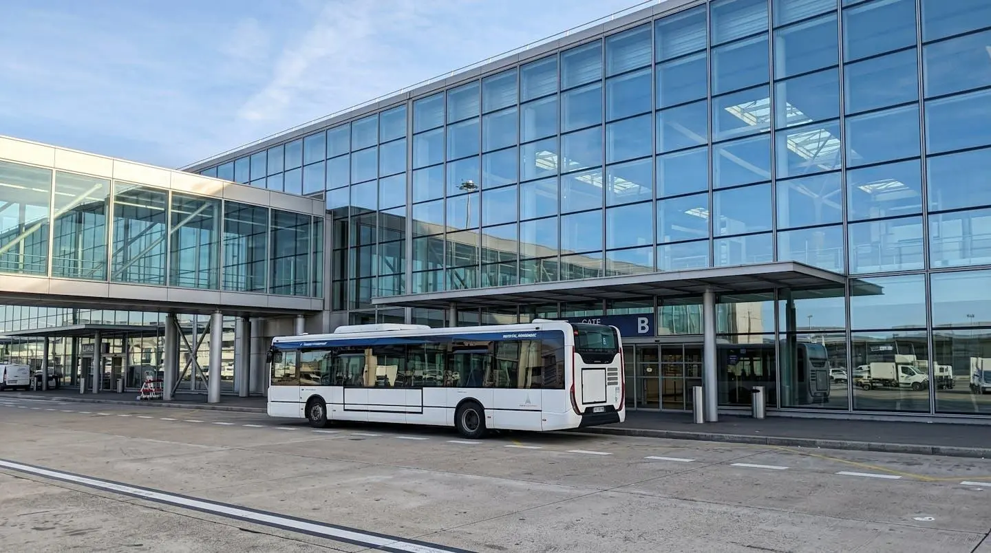 Une navette blanche stationnée devant l'architecture vitrée moderne du terminal de l'aéroport Charles de Gaulle, sans passagers visibles, sous un éclairage naturel de matinée